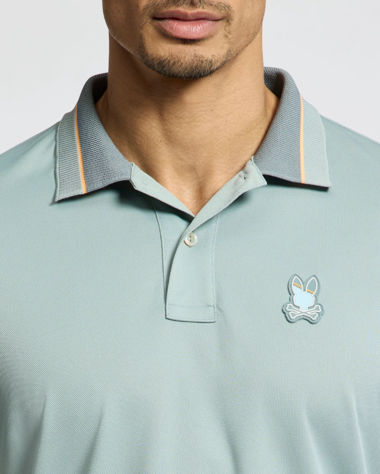 A person is wearing the light green MENS STRATTON SPORT PIQUE POLO by Psycho Bunny, featuring a gray collar, orange trim, and a unique stylized bunny with crossed bones logo. Made from moisture-wicking fabric, it ensures they stay cool and stylish while drawing attention to their attire.