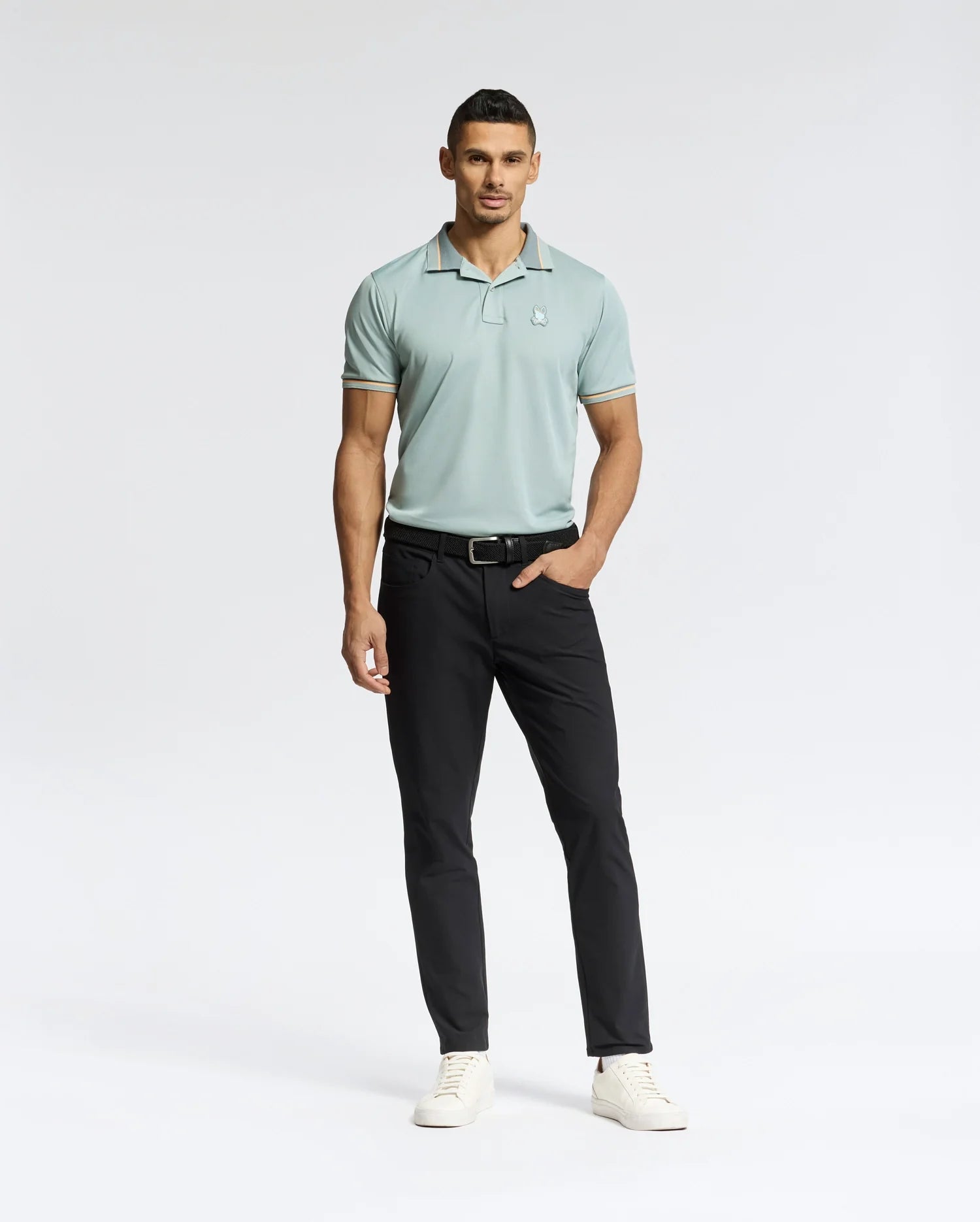 Against a plain white background, a man models in Psycho Bunny's MENS STRATTON SPORT PIQUE POLO - B6K202E200. The light green, moisture-wicking polo pairs with black pants and white sneakers as he stands with his left hand in his pocket, exuding casual sportswear elegance.