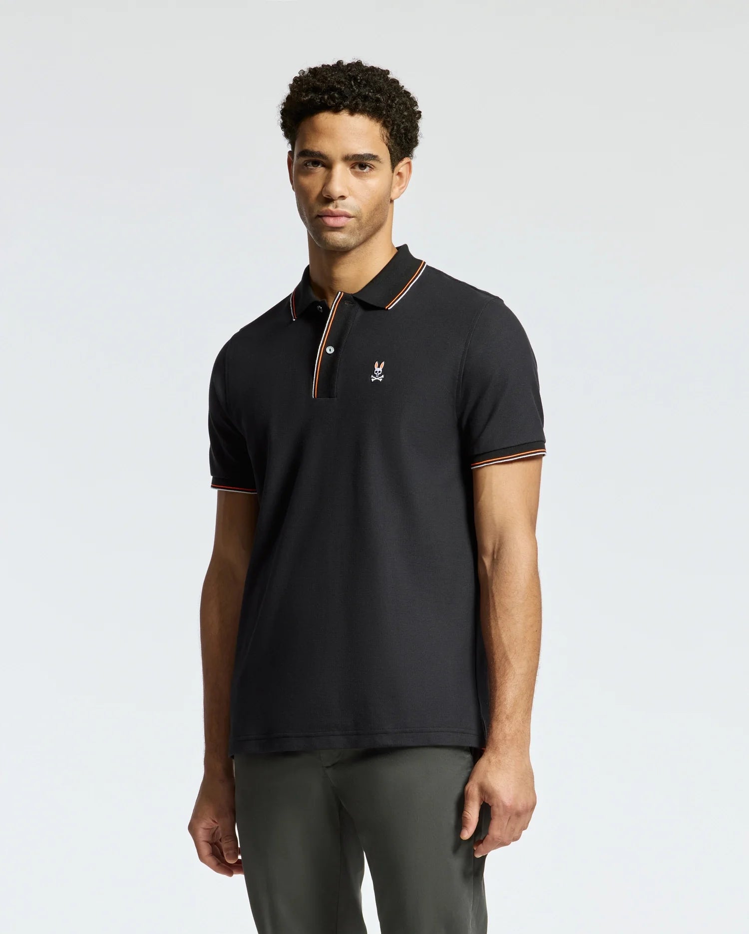 A man wearing the Psycho Bunny MENS BRANDON PIQUE POLO - B6K280E200, distinguished by its black color and orange-striped collar and sleeves, stands against a light gray background. The shirt features a small logo on the left chest. With his short curly hair, he appears relaxed.