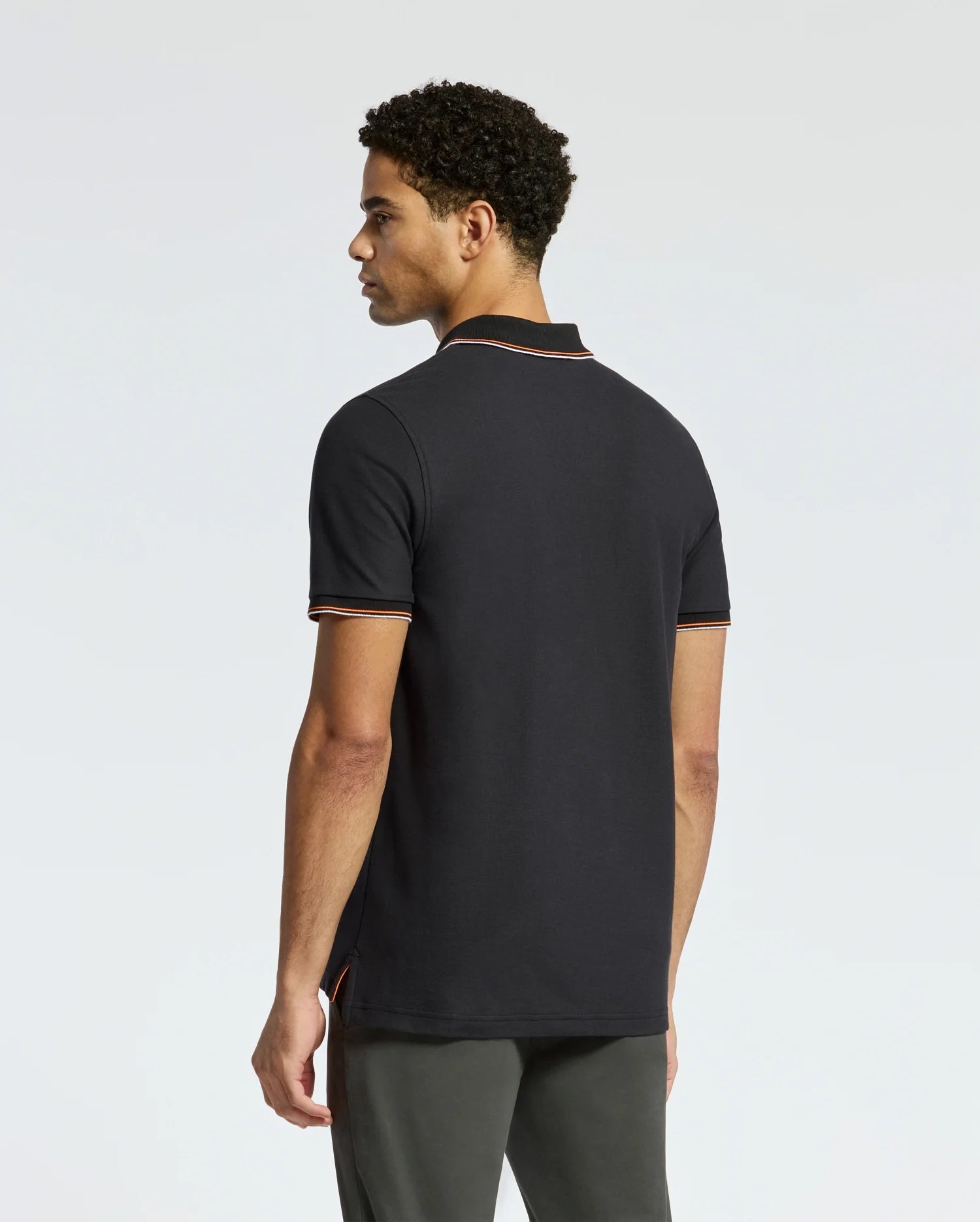 A person wearing the luxurious Psycho Bunny MENS BRANDON PIQUE POLO - B6K280E200, distinguished by its white and orange trim, stands against a plain background. The premium quality shirt showcases short sleeves and a collar. With their dark, curly hair, they are facing slightly away from the camera.