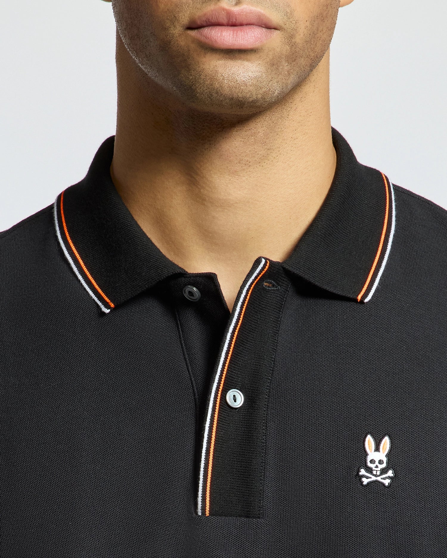 A person is sporting the luxurious Psycho Bunny MENS BRANDON PIQUE POLO - B6K280E200 in black, featuring orange and white trim on the collar. The shirt highlights a small embroidered emblem of a rabbit above a skull and crossbones on the chest, exuding premium quality. The person's face is partially visible.