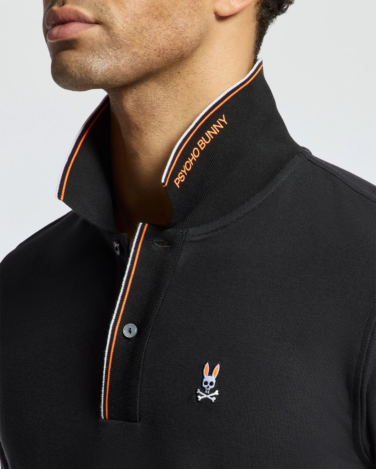 A man is wearing the MENS BRANDON PIQUE POLO - B6K280E200 from Psycho Bunny, which is a black polo shirt featuring an orange and white striped collar. This luxurious men's pique polo showcases a small embroidered bunny with crossbones on the chest and is crafted from premium quality Pima cotton for ultimate comfort.