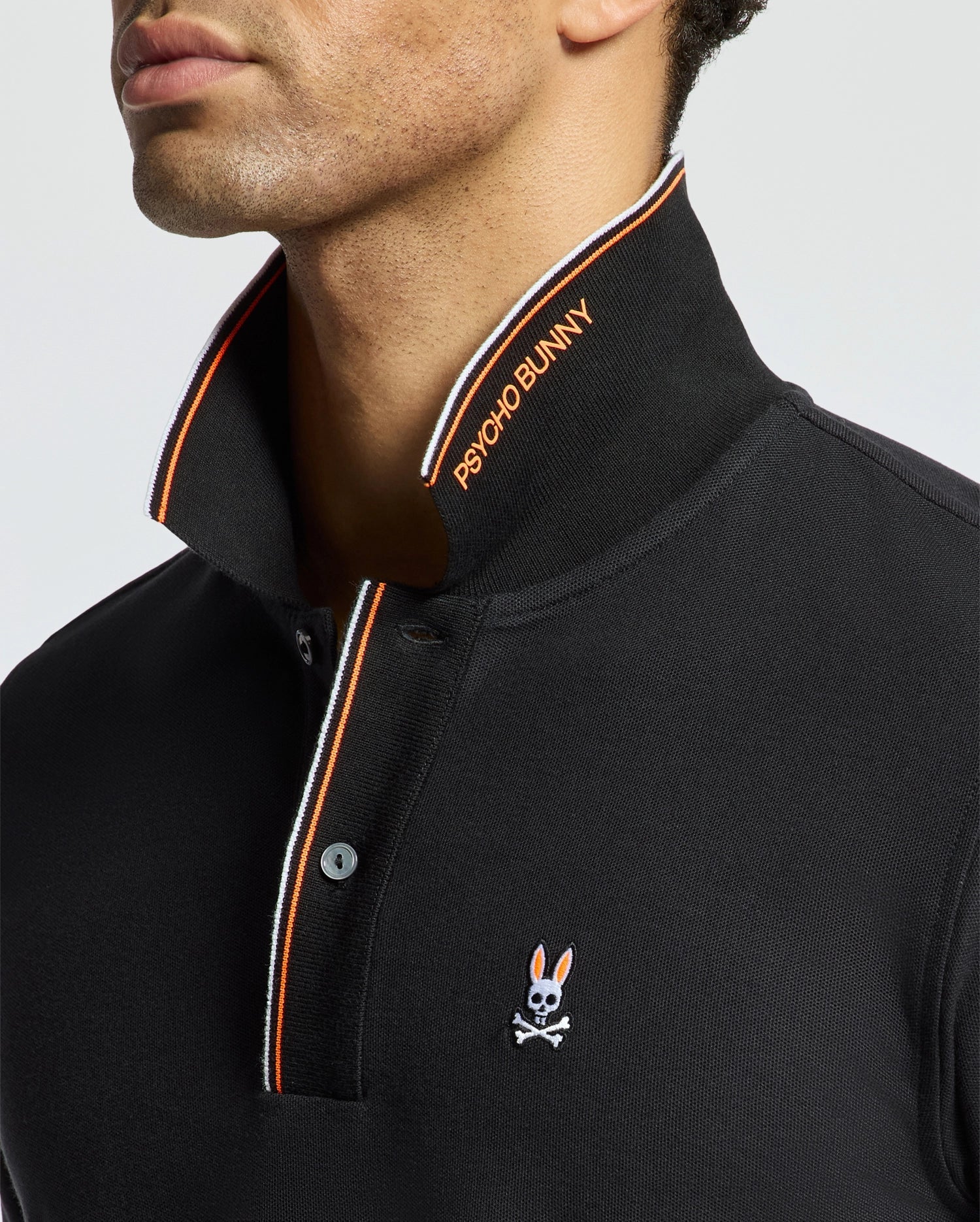 A man is wearing the MENS BRANDON PIQUE POLO - B6K280E200 from Psycho Bunny, which is a black polo shirt featuring an orange and white striped collar. This luxurious men's pique polo showcases a small embroidered bunny with crossbones on the chest and is crafted from premium quality Pima cotton for ultimate comfort.