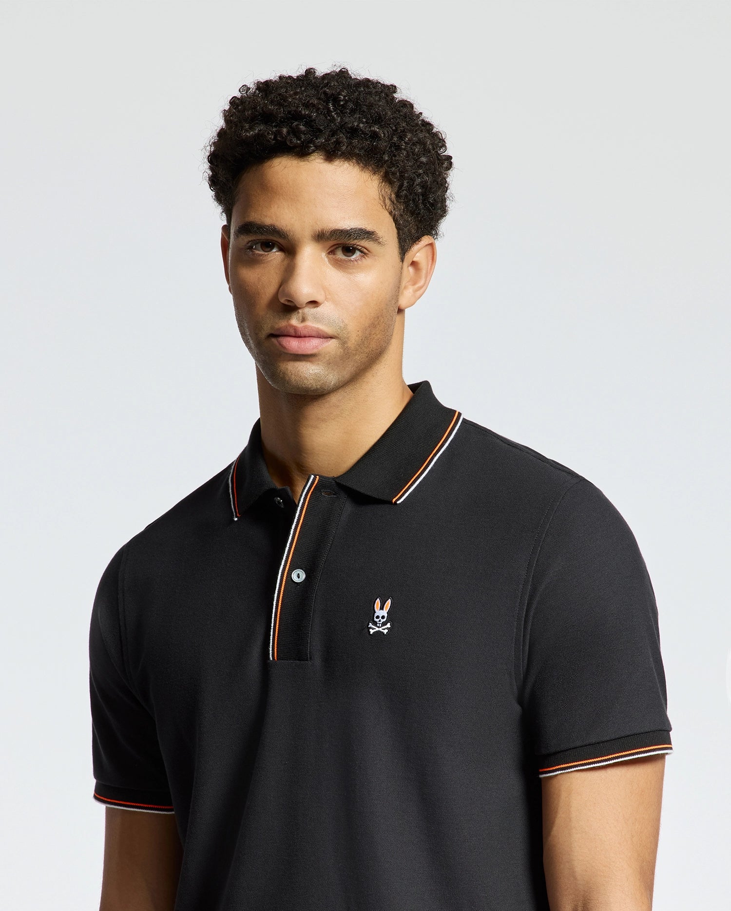 A man wearing the luxurious Psycho Bunny MENS BRANDON PIQUE POLO (B6K280E200) with a discreet embroidered logo on the chest stands against a plain background. This premium quality polo is distinguished by striped detailing on the collar and sleeves, enhancing its elegance.
