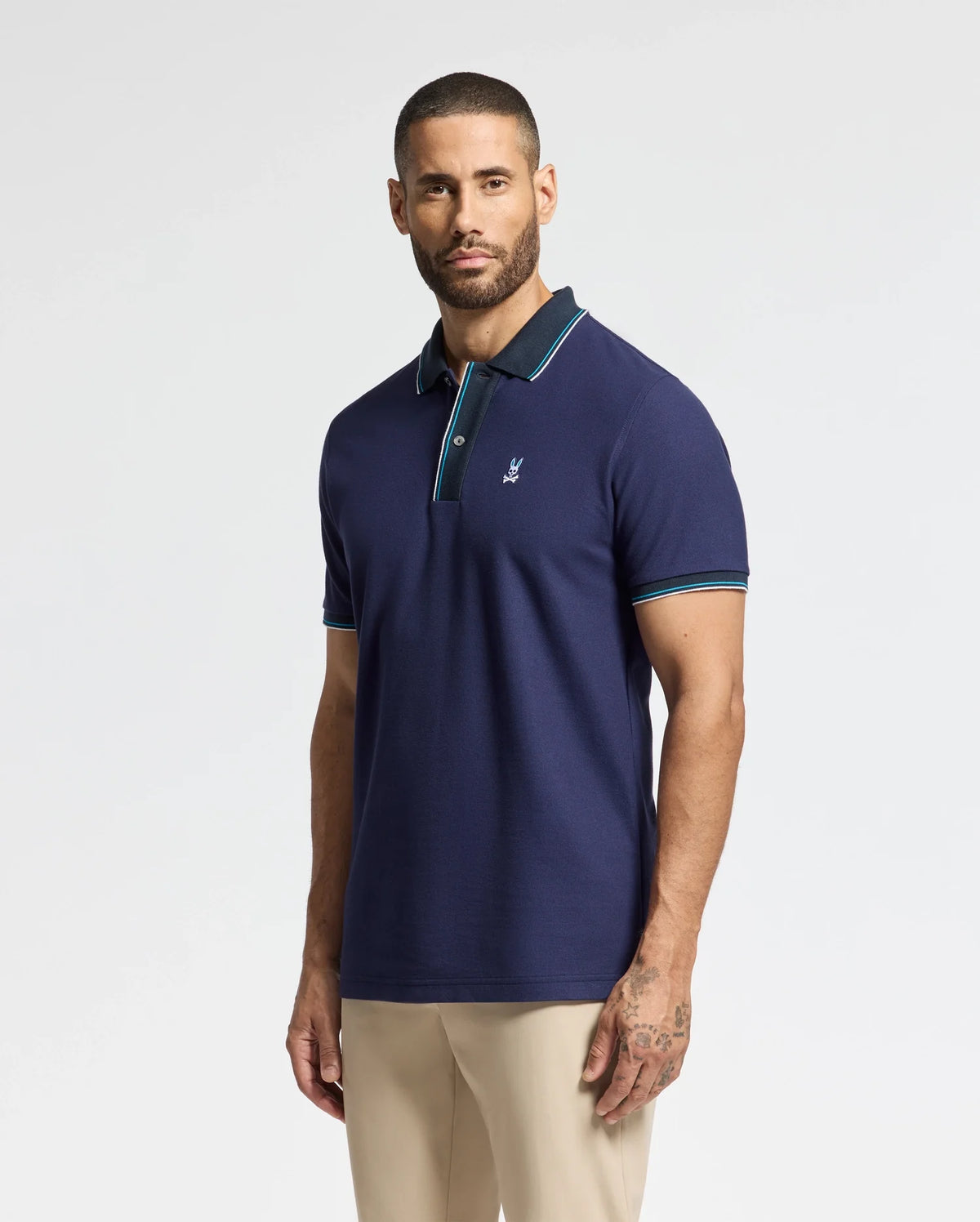 A man with short hair and a beard is wearing the Psycho Bunny MENS BRANDON PIQUE POLO in navy blue, featuring green and white trim. He pairs it with beige pants while standing against a plain background, revealing visible tattoos on his right arm.