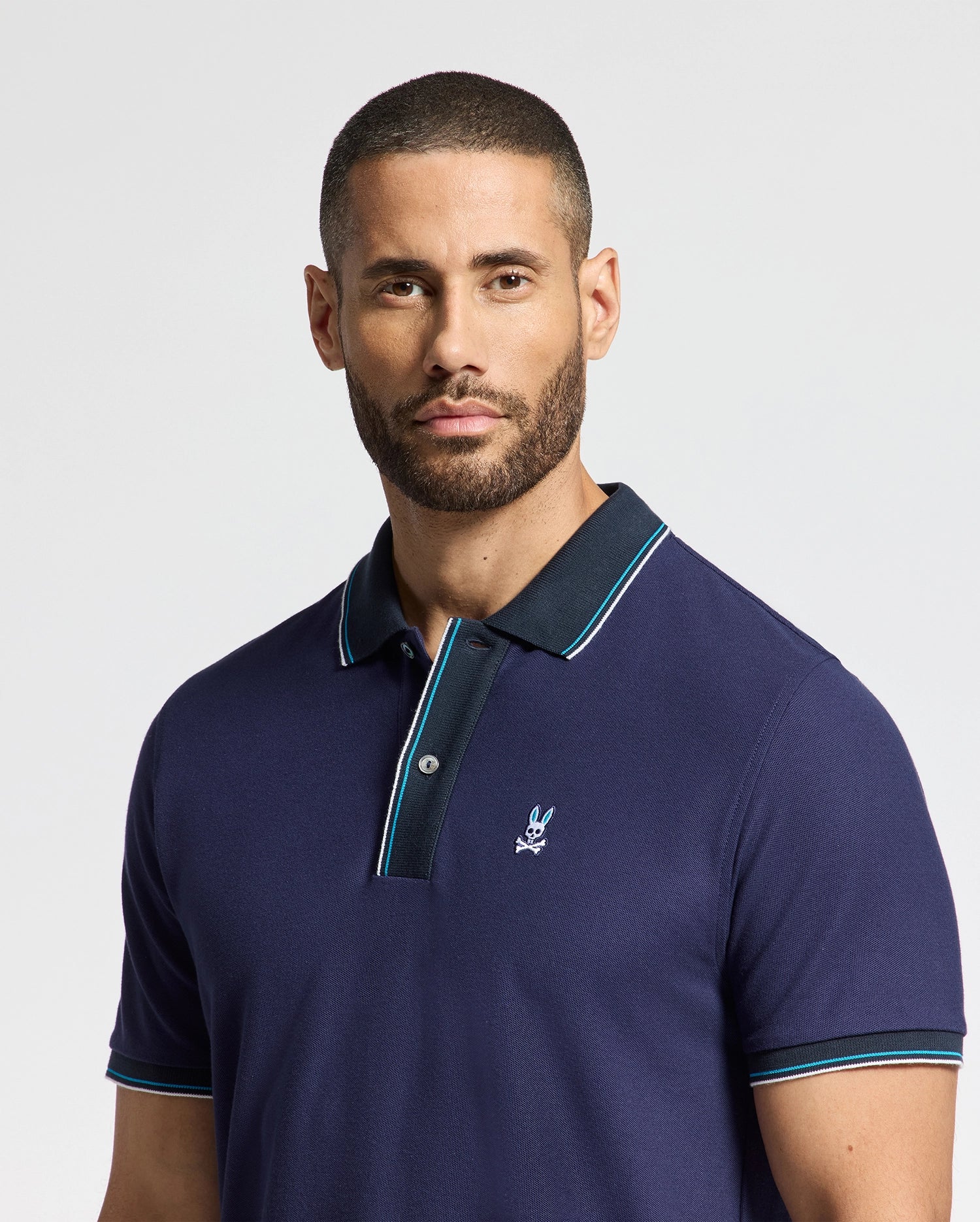 A man with short hair and a beard wears the Psycho Bunny MENS BRANDON PIQUE POLO - B6K280E200, crafted from luxurious cotton pique in navy blue and featuring a logo on the chest. The polo's black collar and light blue trim enhance its elegance as he gazes directly at the camera against a light gray backdrop.