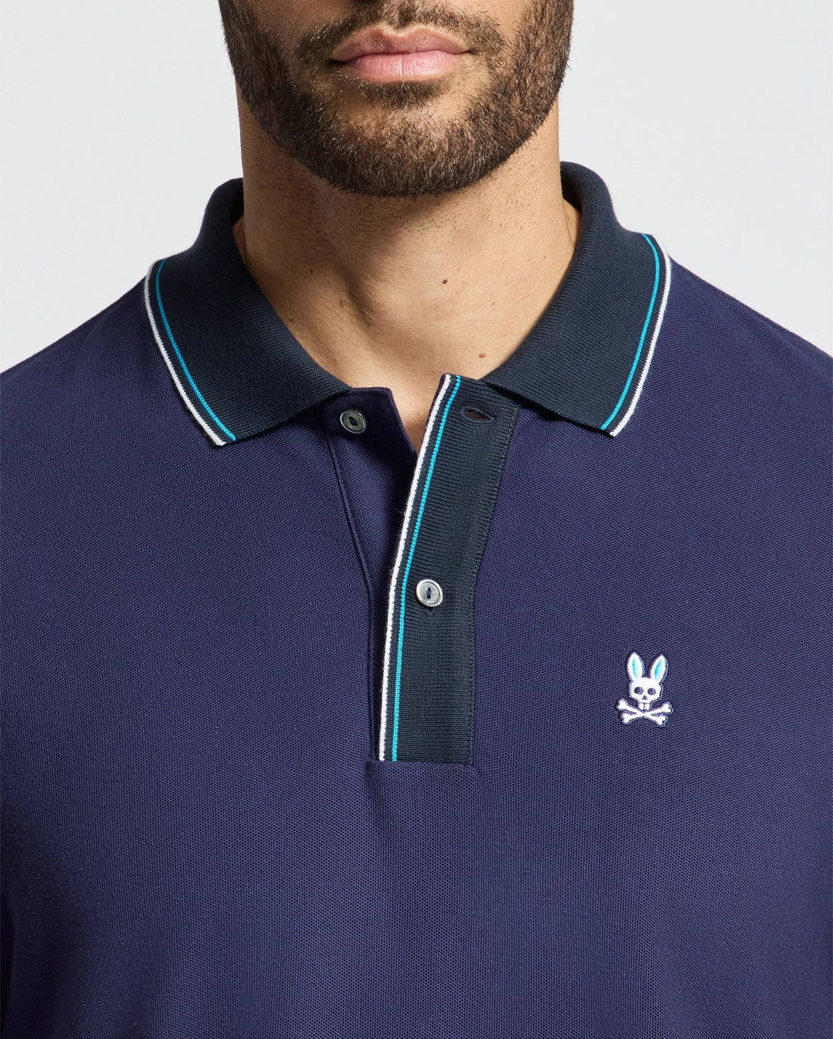 A man wears the Psycho Bunny luxurious MENS BRANDON PIQUE POLO - B6K280E200 in navy blue, accented with white and light blue trim on the collar and sleeves. This polo shirt of premium quality is adorned with an embroidered rabbit with crossed bones on the chest. The soft hues complement a neutral light gray background seamlessly.