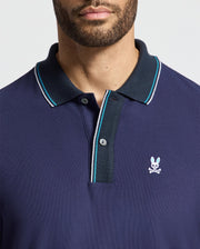 A man wears the Psycho Bunny luxurious MENS BRANDON PIQUE POLO - B6K280E200 in navy blue, accented with white and light blue trim on the collar and sleeves. This polo shirt of premium quality is adorned with an embroidered rabbit with crossed bones on the chest. The soft hues complement a neutral light gray background seamlessly.