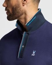 A person wearing a navy blue MENS BRANDON PIQUE POLO - B6K280E200, designed by Psycho Bunny, adorned with a white and teal trim. The collar is turned up, showcasing the "Psycho Bunny" logo. This premium quality polo shirt also features a small emblem of a bunny with crossed bones on the chest.