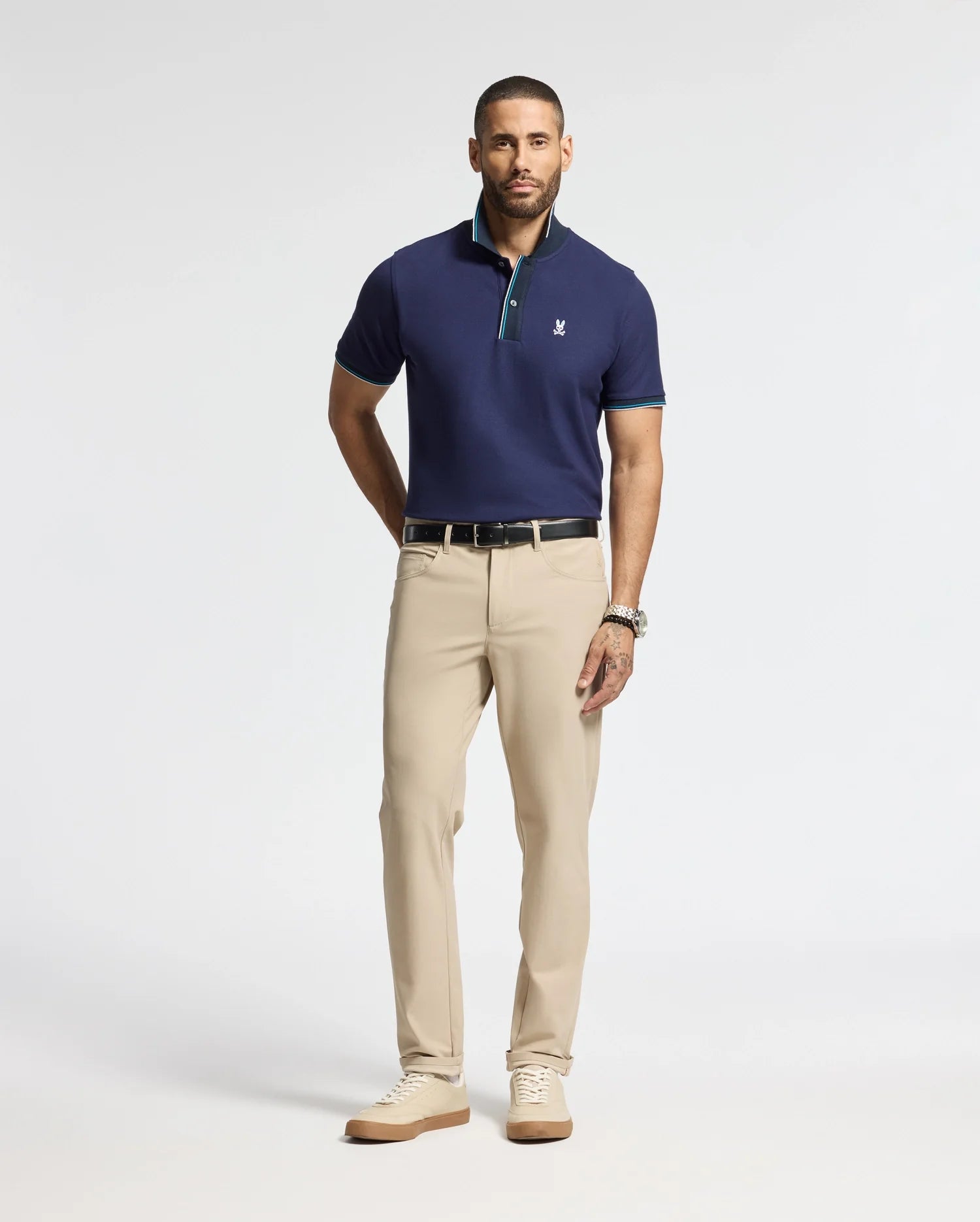 A man stands against a plain background, wearing a Psycho Bunny MENS BRANDON PIQUE POLO in navy blue, featuring a small logo on the chest. He pairs it with beige trousers, a black belt, tan shoes, and a wristwatch. He has short hair and a trimmed beard.