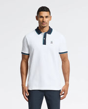A man confidently stands against a plain white background wearing the Psycho Bunny MENS BRANDON PIQUE POLO - B6K280E200, a luxurious pique polo in white with black and blue trim. The small logo on the chest complements his short dark hair, highlighting the premium quality of this piece.