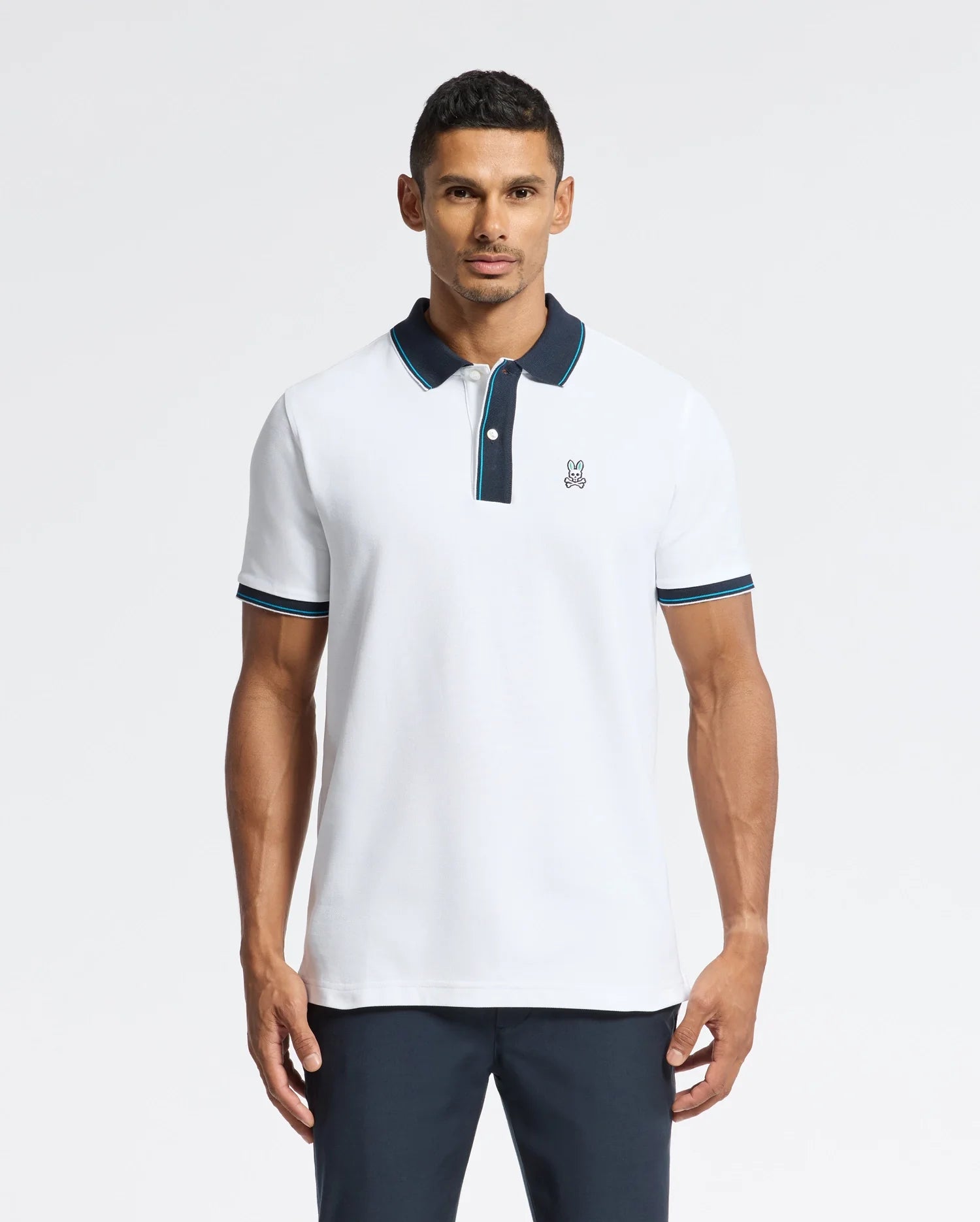 A man confidently stands against a plain white background wearing the Psycho Bunny MENS BRANDON PIQUE POLO - B6K280E200, a luxurious pique polo in white with black and blue trim. The small logo on the chest complements his short dark hair, highlighting the premium quality of this piece.