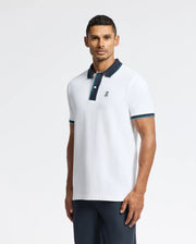A man wearing a luxurious Psycho Bunny MENS BRANDON PIQUE POLO - B6K280E200, featuring a dark blue collar and sleeve trim, stands against a plain white background. The premium quality polo shirt is adorned with a small embroidered logo on the chest. He looks straight ahead with his arms at his sides, exuding effortless style.