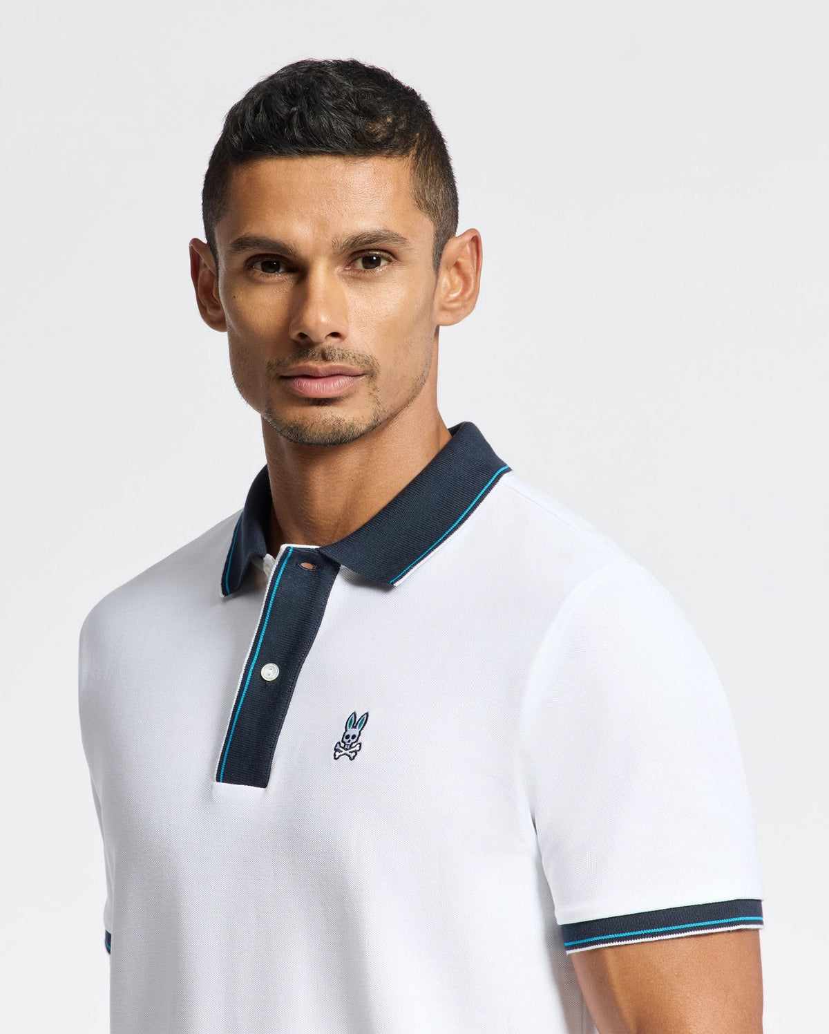 A man is dressed in the stylish MENS BRANDON PIQUE POLO by Psycho Bunny, featuring a dark collar and a discreet embroidered logo on the chest. He has short dark hair and a well-maintained beard, posing against a simple light background.