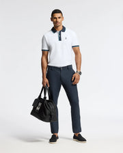 A man poses in a premium quality MENS BRANDON PIQUE POLO (SKU: B6K280E200) from Psycho Bunny and dark trousers, holding a black bag. He wears black shoes and a watch, standing against a plain white background.