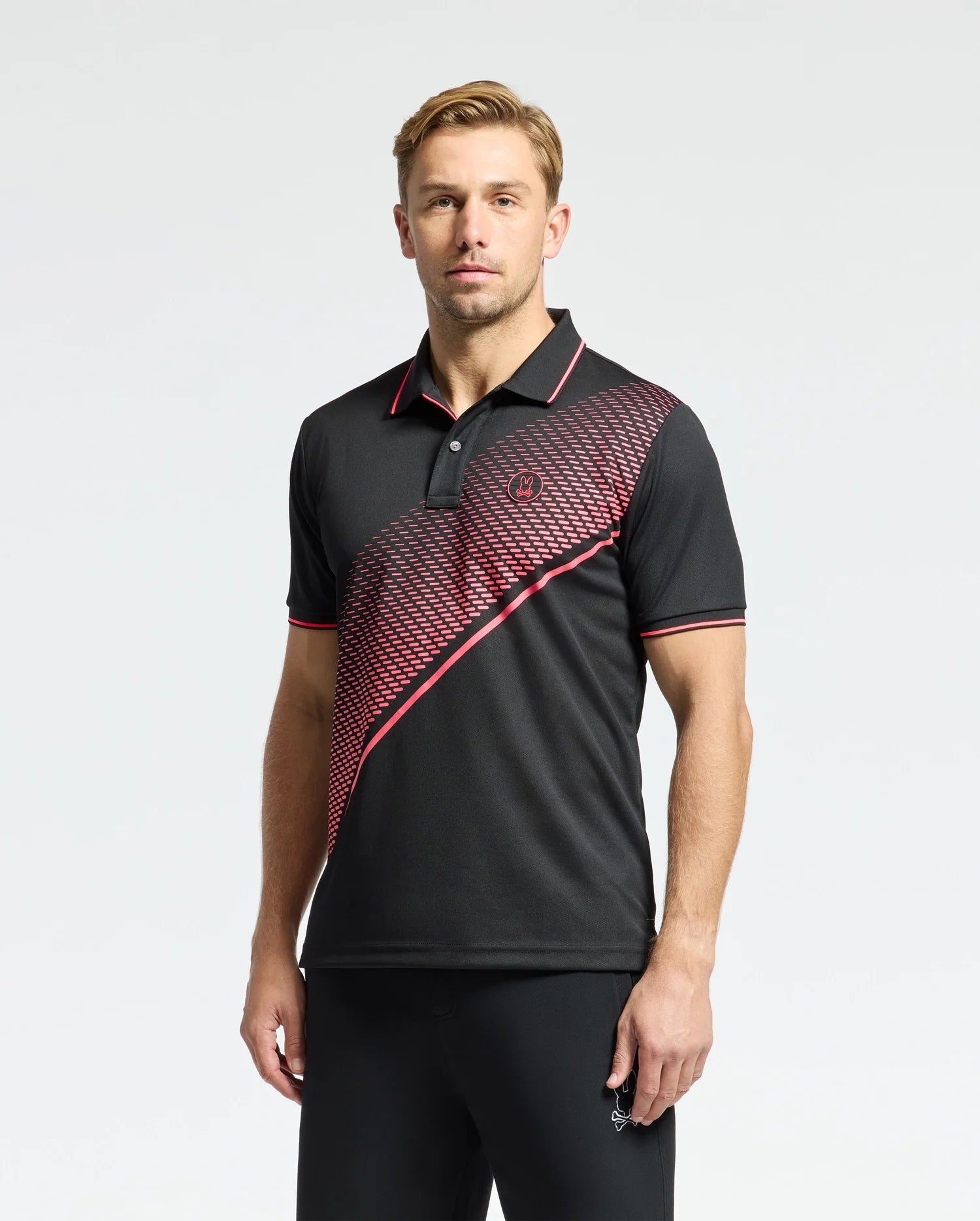 A man with short light brown hair poses against a plain background, wearing the Psycho Bunny MENS MACK SPORT PIQUE POLO (B6K321E200), a black shirt with red accents and a diagonal red patterned stripe. Crafted for comfort with Peruvian skill, it features moisture-wicking technology.