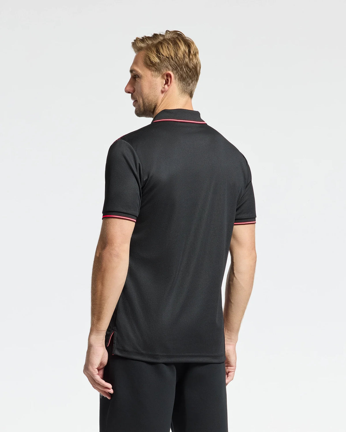 A man with short blond hair, facing away, wears Psycho Bunny's MENS MACK SPORT PIQUE POLO (B6K321E200) in black with red trim and black shorts. Engineered with moisture-wicking technology, his outfit ensures he stays cool against the plain white background.