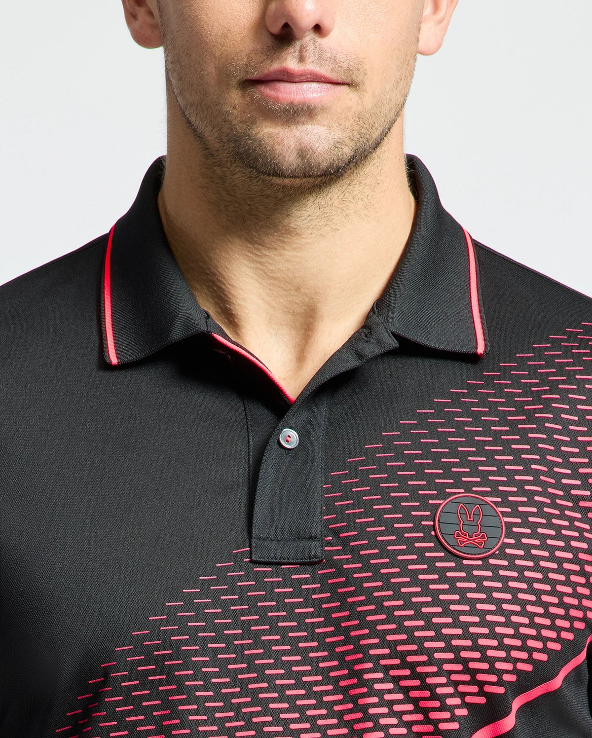 A person is seen wearing the Psycho Bunny MENS MACK SPORT PIQUE POLO - B6K321E200, featuring a black color with red accents and a patterned design on the right side. The quick-dry shirt showcases Peruvian craftsmanship and a rabbit logo with crossed ears, while only their lower face is shown.