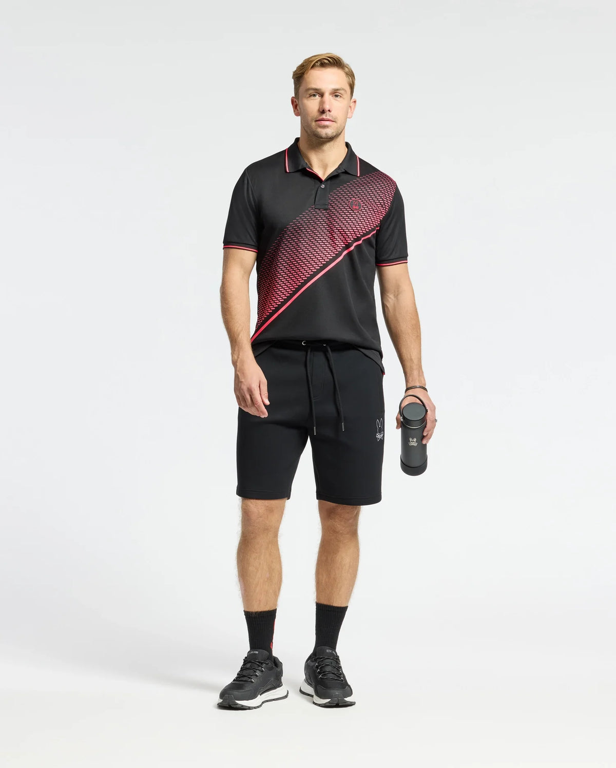 A man in a Psycho Bunny MENS MACK SPORT PIQUE POLO - B6K321E200, featuring moisture-wicking, black with red diagonal stripes, stands with a water bottle and matching shorts. He wears black sneakers and socks, facing the camera against a plain white background.