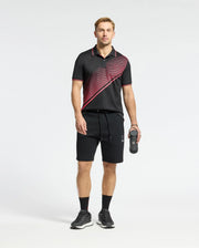 A man in a Psycho Bunny MENS MACK SPORT PIQUE POLO - B6K321E200, featuring moisture-wicking, black with red diagonal stripes, stands with a water bottle and matching shorts. He wears black sneakers and socks, facing the camera against a plain white background.