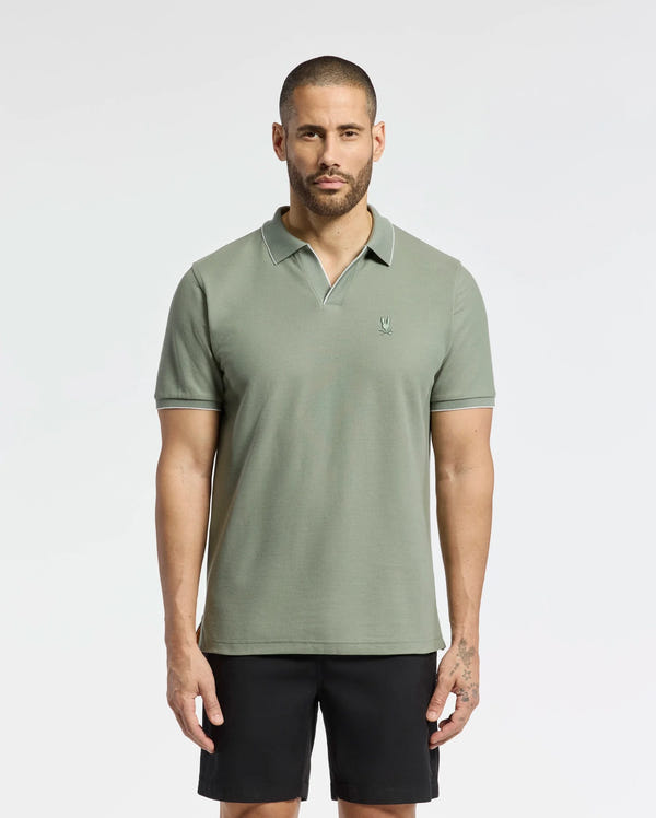 A man with a buzz cut in the Psycho Bunny MENS EAST HILLS JOHNNY COLLAR POLO in light olive green and black shorts stands against a white background. The 100% Pima cotton shirt features an embroidered logo on the left chest, offering a touch of relaxed sophistication.