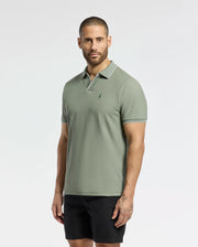 A bearded man with short hair wears Psycho Bunny's MENS EAST HILLS JOHNNY COLLAR POLO - B6K331B200 in sage green, made of 100% Pima cotton with a chest emblem. Paired with black shorts, he stands against a plain white background, exuding a subtle seaside vibe.