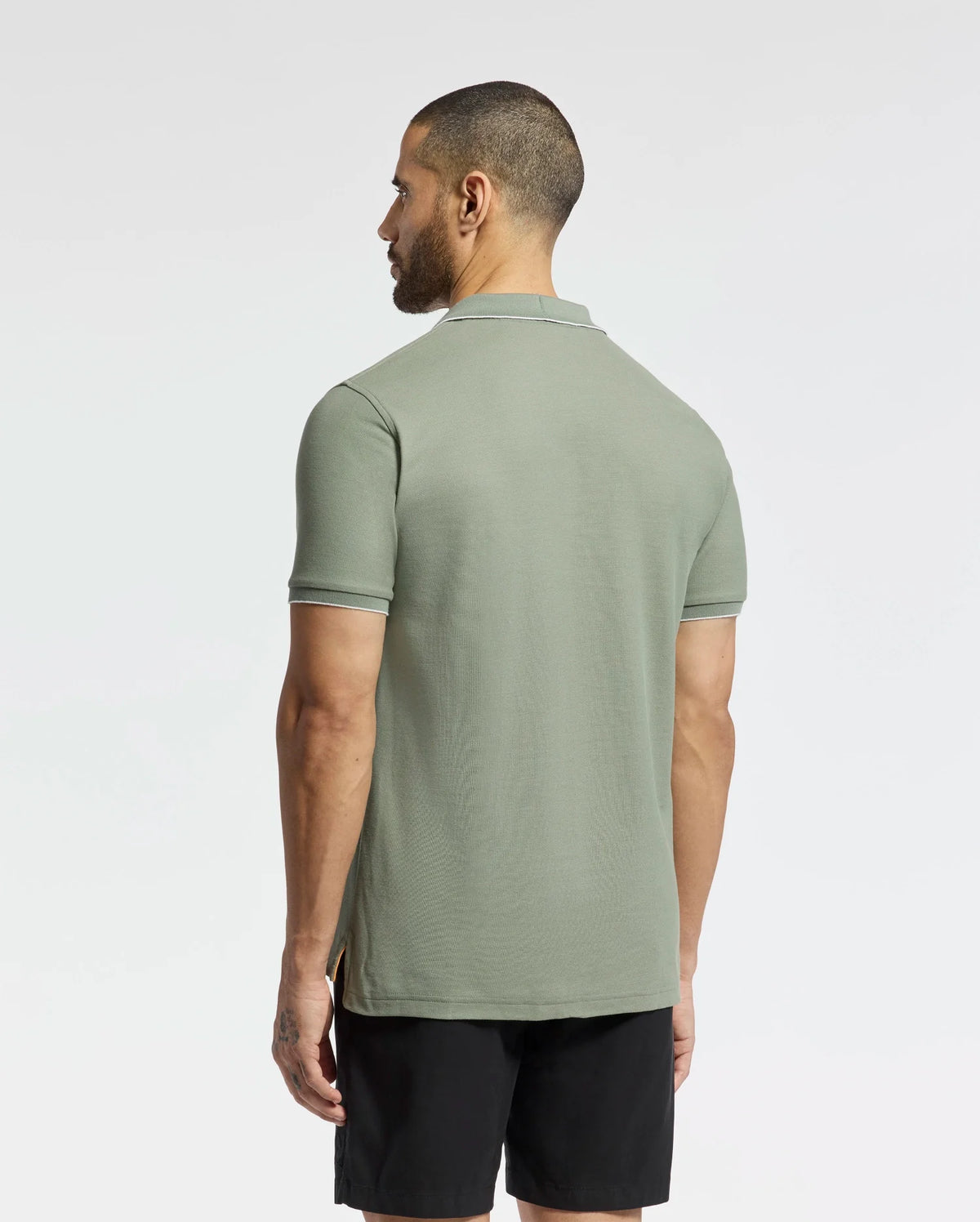 A man with short hair stands facing away, wearing the Psycho Bunny East Hills Johnny Collar Polo in light green with white-trimmed sleeves, made from 100% Pima cotton. His black shorts complement the seaside vibe against a plain white background.