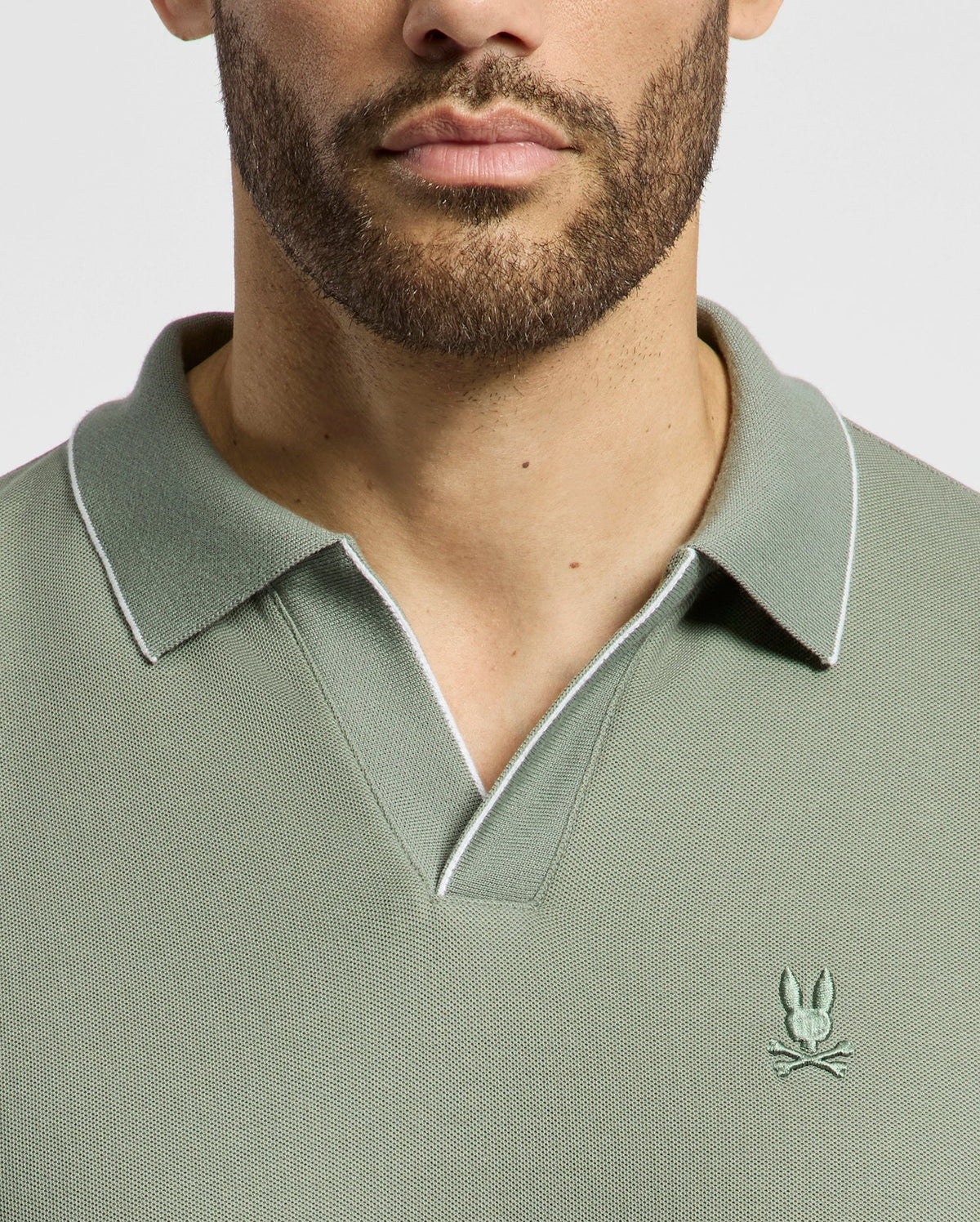 The image shows a person wearing Psycho Bunny's MENS EAST HILLS JOHNNY COLLAR POLO in sage green, made from 100% Pima cotton. A small embroidered rabbit over crossed bones adds flair, while the Johnny collar enhances its seaside vibe against a plain white background.