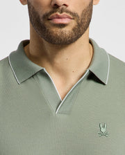 The image shows a person wearing Psycho Bunny's MENS EAST HILLS JOHNNY COLLAR POLO in sage green, made from 100% Pima cotton. A small embroidered rabbit over crossed bones adds flair, while the Johnny collar enhances its seaside vibe against a plain white background.