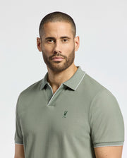 A man with a neatly trimmed beard wears Psycho Bunny's MENS EAST HILLS JOHNNY COLLAR POLO (B6K331B200), a light green, 100% Pima cotton polo featuring an embroidered chest logo. He stands against a plain, light backdrop with a neutral expression, embodying coastal elegance.