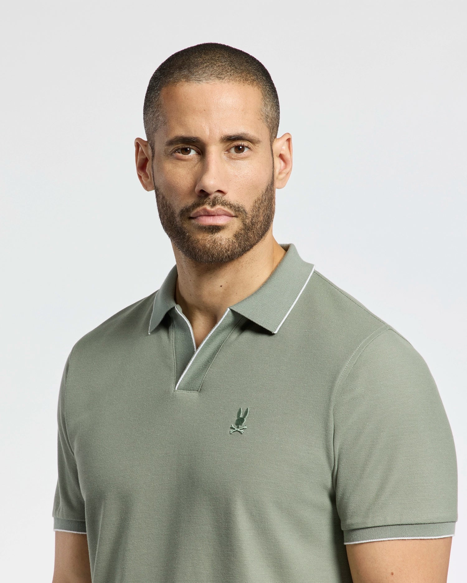 A man with a neatly trimmed beard wears Psycho Bunny's MENS EAST HILLS JOHNNY COLLAR POLO (B6K331B200), a light green, 100% Pima cotton polo featuring an embroidered chest logo. He stands against a plain, light backdrop with a neutral expression, embodying coastal elegance.