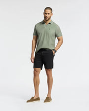 A man stands against a plain white background, wearing a Psycho Bunny MENS EAST HILLS JOHNNY COLLAR POLO in light green and black shorts, paired with brown loafers. Crafted from 100% Pima cotton, his look radiates a subtle seaside vibe. He has short hair and a beard, showcasing a watch on his left wrist.