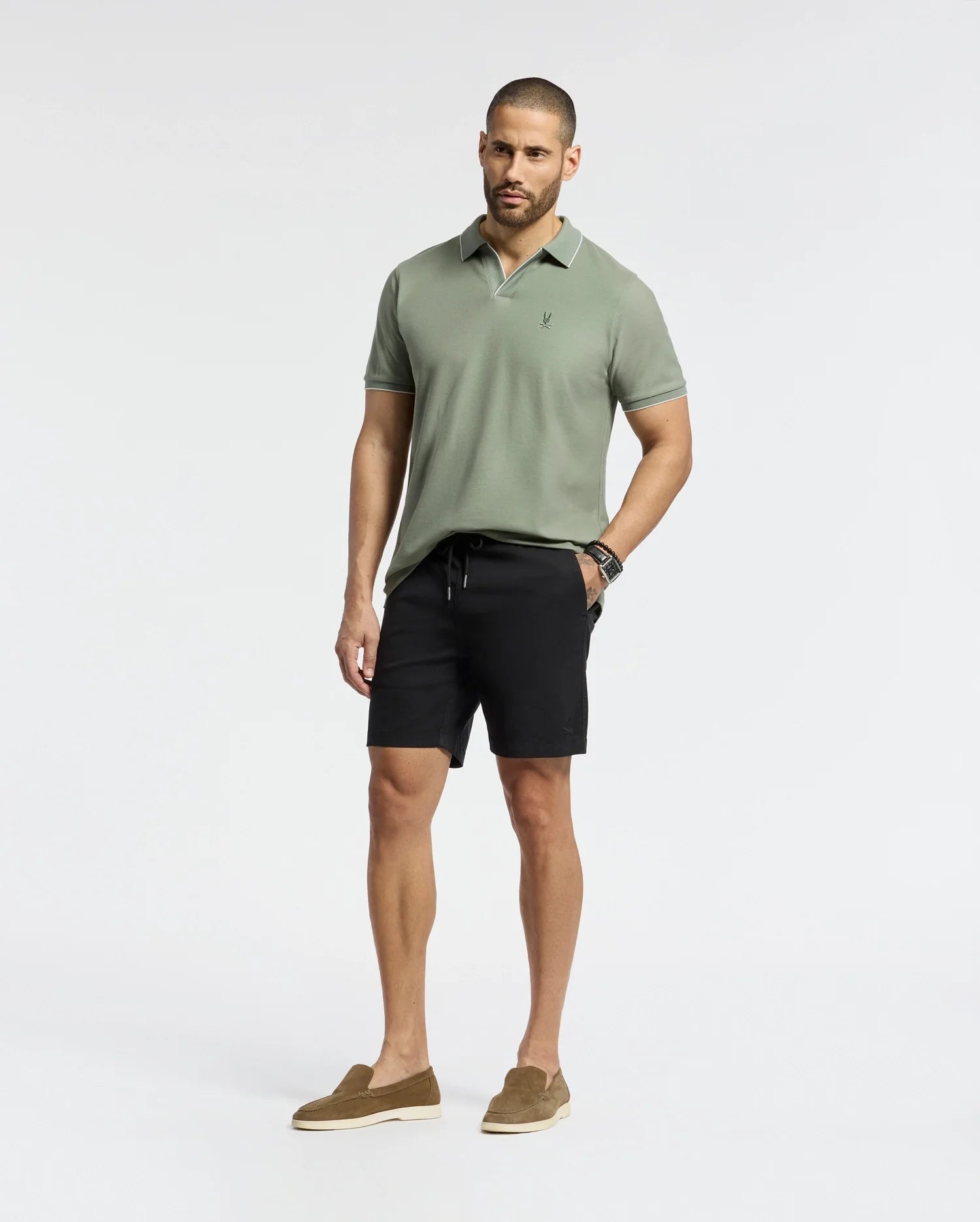 A man stands against a plain white background, wearing a Psycho Bunny MENS EAST HILLS JOHNNY COLLAR POLO in light green and black shorts, paired with brown loafers. Crafted from 100% Pima cotton, his look radiates a subtle seaside vibe. He has short hair and a beard, showcasing a watch on his left wrist.