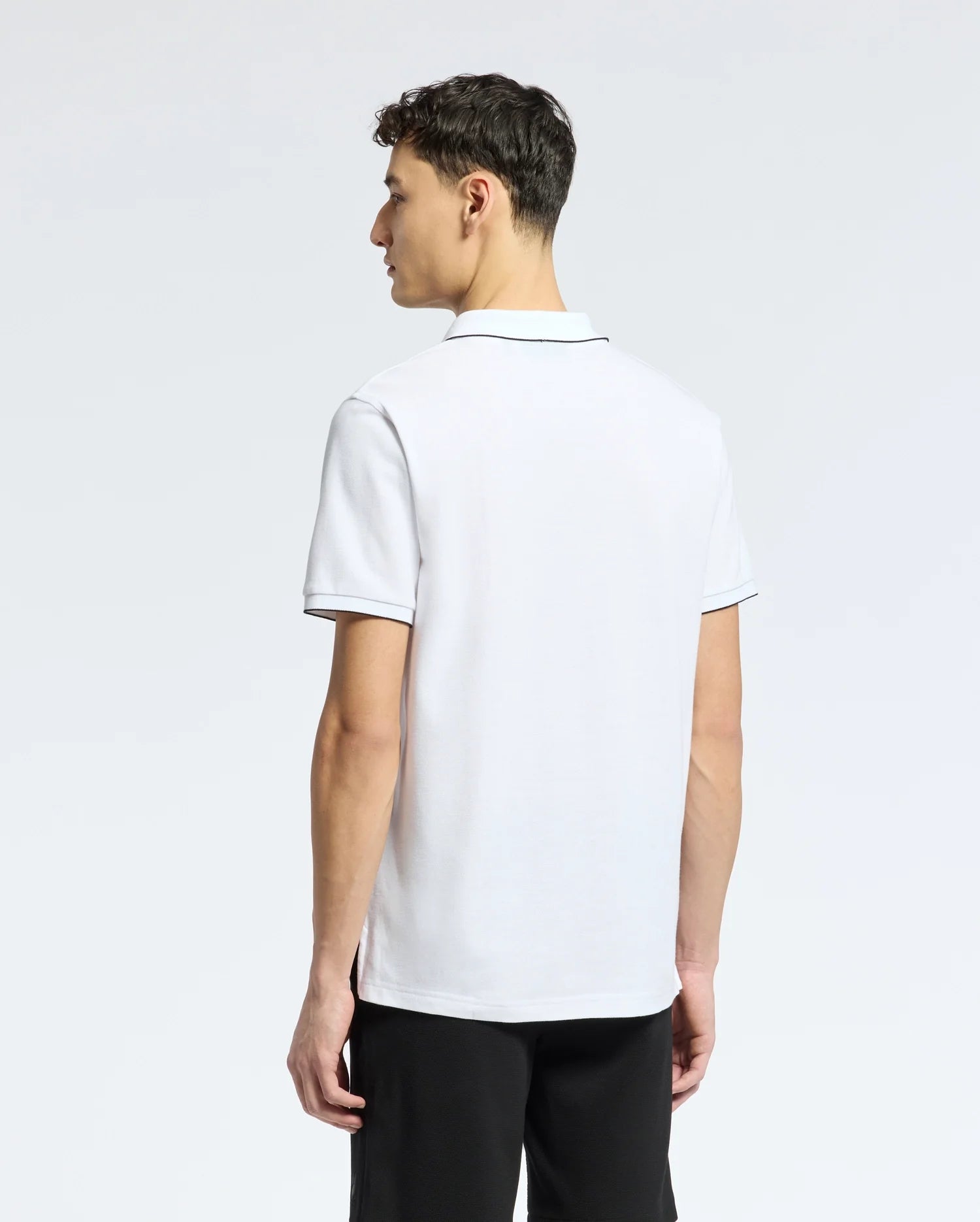 Facing away, a person wears the Psycho Bunny Mens East Hills Johnny Collar Polo (B6K331B200) in white with black trim. The background is plain and light-colored. They have short, dark hair and are wearing black pants.