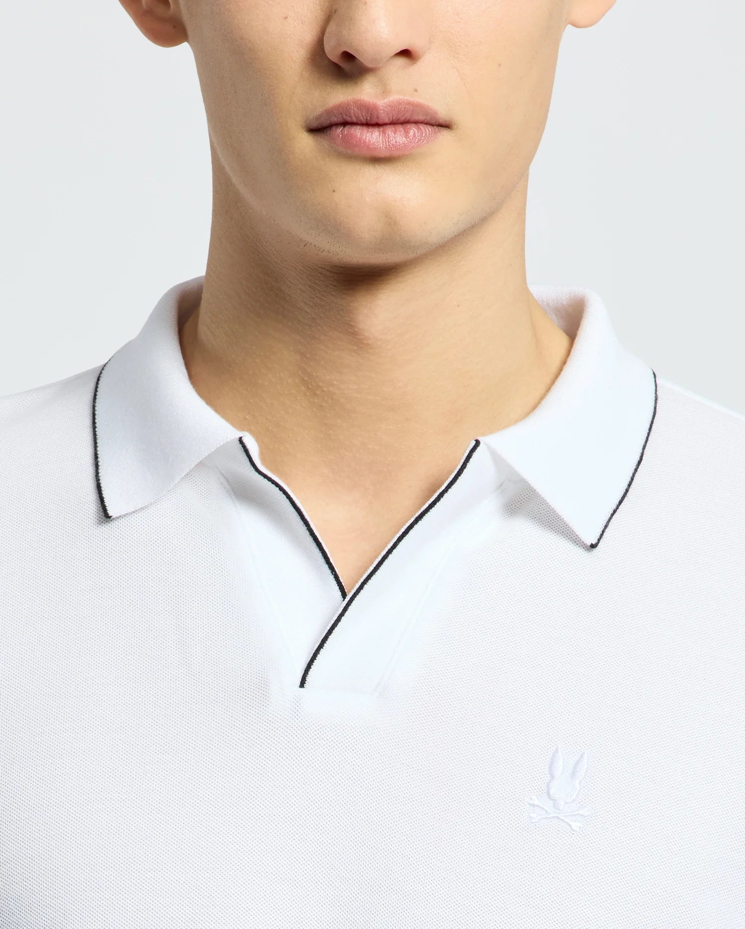 A person wears a white Psycho Bunny East Hills Johnny Collar Polo (B6K331B200) made of luxurious Pima cotton, featuring a small embroidered logo on the chest and dark-trimmed collar, with focus on the upper torso and part of the face against a plain background.