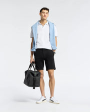 A person is dressed in a Psycho Bunny MENS EAST HILLS JOHNNY COLLAR POLO - B6K331B200, complemented by a light blue sweater draped over the shoulders. They wear black shorts and white sneakers, holding a black duffel bag, all set against a plain white background.