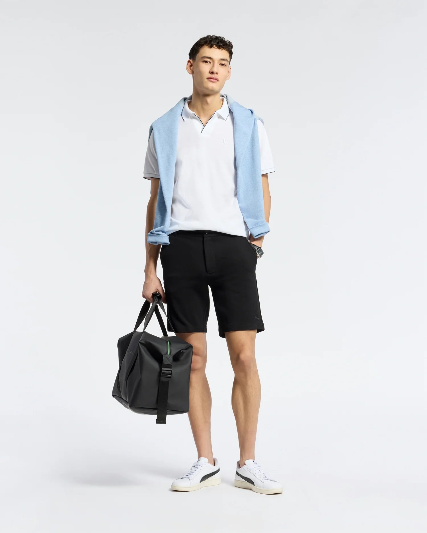 A person is dressed in a Psycho Bunny MENS EAST HILLS JOHNNY COLLAR POLO - B6K331B200, complemented by a light blue sweater draped over the shoulders. They wear black shorts and white sneakers, holding a black duffel bag, all set against a plain white background.