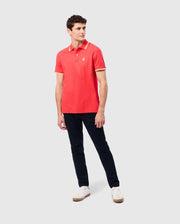 A man is standing against a plain background wearing the Psycho Bunny MENS GRANBURY PIQUE POLO (B6K400C200), which features a tipping collar in red and yellow trim, paired with dark blue pants and white sneakers. His left arm is relaxed by his side while his right arm is slightly bent at the elbow. He is looking to his right.