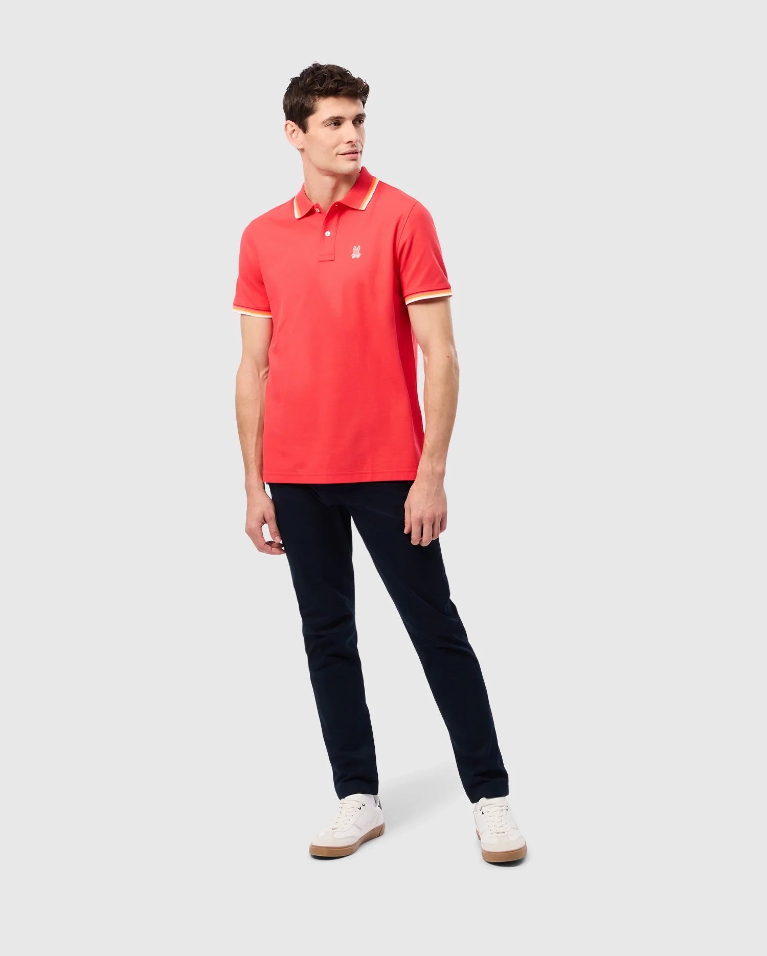 A man is standing against a plain background wearing the Psycho Bunny MENS GRANBURY PIQUE POLO (B6K400C200), which features a tipping collar in red and yellow trim, paired with dark blue pants and white sneakers. His left arm is relaxed by his side while his right arm is slightly bent at the elbow. He is looking to his right.