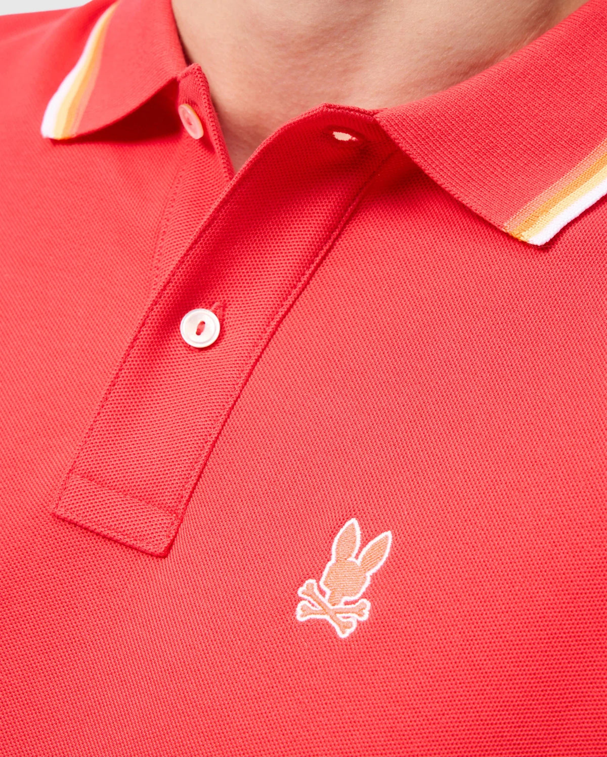A close-up image of a person wearing the MENS GRANBURY PIQUE POLO - B6K400C200 by Psycho Bunny, showcasing its red Pima cotton fabric with white and yellow striped rib knit trim on the collar. The polo shirt features a small embroidered logo of a rabbit head with crossbones just below the buttons.