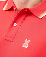 A close-up image of a person wearing the MENS GRANBURY PIQUE POLO - B6K400C200 by Psycho Bunny, showcasing its red Pima cotton fabric with white and yellow striped rib knit trim on the collar. The polo shirt features a small embroidered logo of a rabbit head with crossbones just below the buttons.