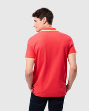 A person with short dark hair is standing and facing away from the camera. They are wearing a bright red Psycho Bunny MENS GRANBURY PIQUE POLO - B6K400C200 with white trim on the sleeves and collar. Their hands are in the pockets of their dark pants, and the background is light gray.