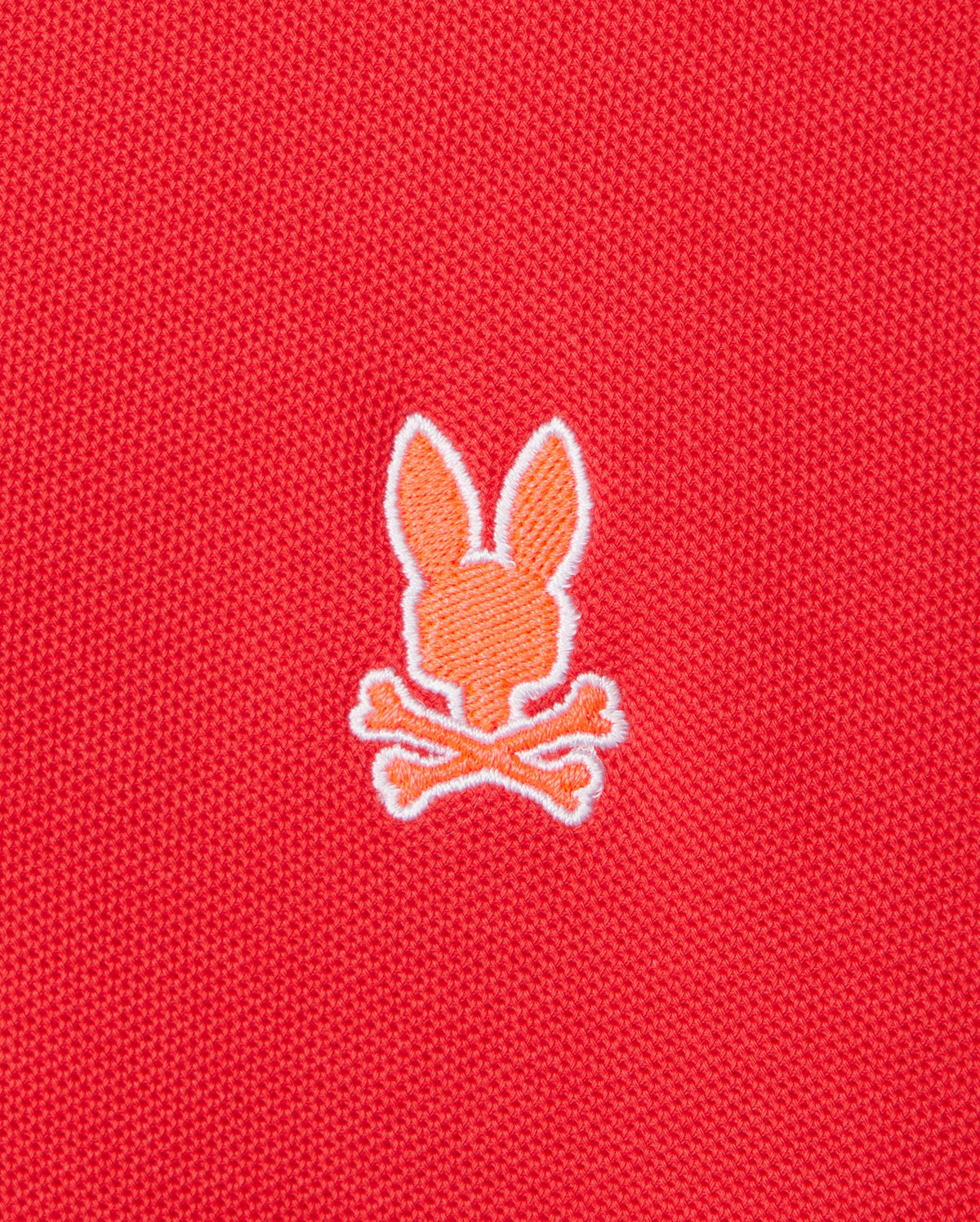 An embroidered orange silhouette of a bunny head with two ears, positioned above a pair of crossed bones in a skull and crossbones motif, adorns the front of the red Pima cotton "MENS GRANBURY PIQUE POLO - B6K400C200" by Psycho Bunny.