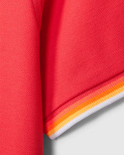 Close-up of the sleeve of the MENS GRANBURY PIQUE POLO - B6K400C200 by Psycho Bunny. The end of the red sports jersey sleeve is detailed with thin yellow and orange stripes, along with a slightly larger white stripe at the edge. The textured fabric, reminiscent of diamond-knit Pima cotton piqué, appears breathable.