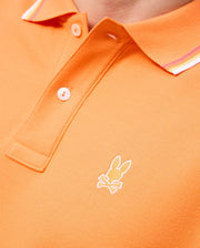 Close-up of an orange MENS GRANBURY PIQUE POLO - B6K400C200 by Psycho Bunny crafted from diamond-knit Pima cotton, featuring white buttons and a tipping collar with red and white striped accents. The shirt has an embroidered logo of a rabbit's head with crossbones below it on the chest.