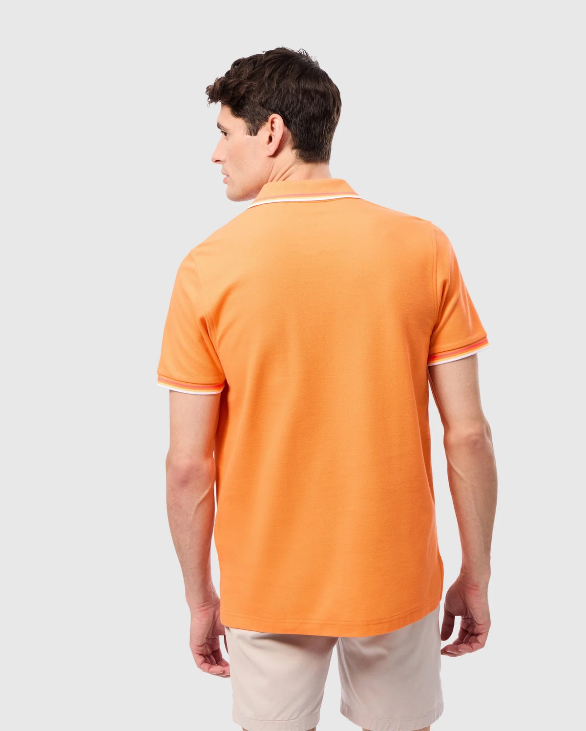 A man with short brown hair is standing and facing away from the camera. He dons a bright orange Psycho Bunny MENS GRANBURY PIQUE POLO - B6K400C200 with white trim tipping the collar and sleeves, paired with beige shorts. The shirt's diamond-knit Pima cotton fabric appears comfortable against the plain light gray background.