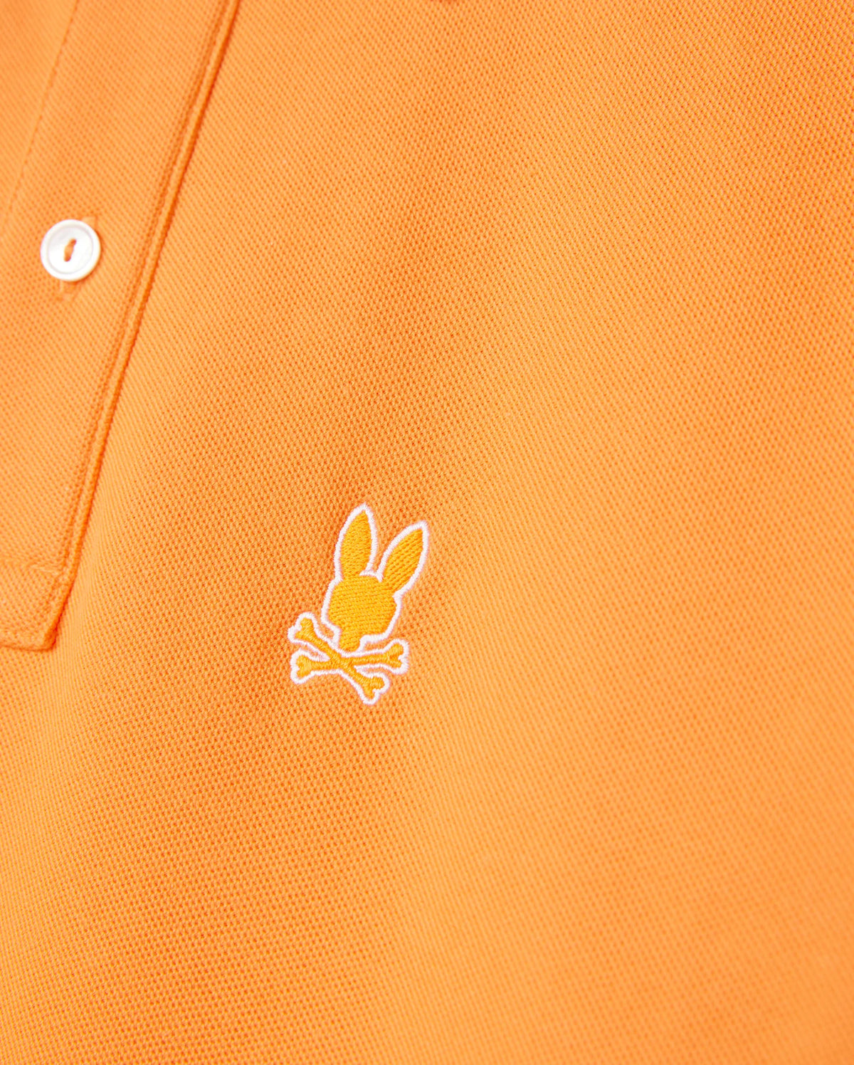 Close-up of an orange MENS GRANBURY PIQUE POLO - B6K400C200 by Psycho Bunny featuring a white button on the placket. The shirt has an embroidered logo of a stylized rabbit's head with crossed bones underneath, located on the left chest area. Made from diamond-knit Pima cotton, the fabric appears to be both textured and durable.