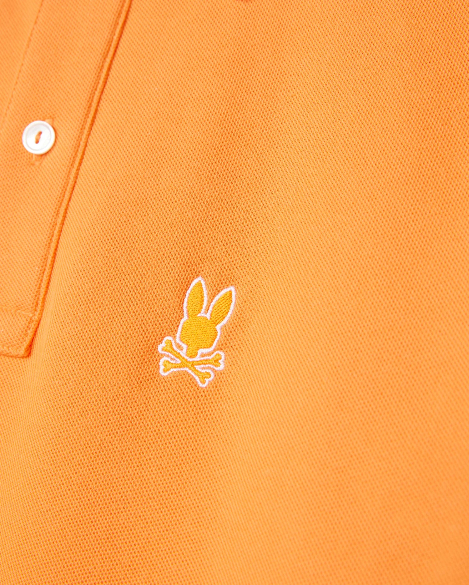 Close-up of an orange MENS GRANBURY PIQUE POLO - B6K400C200 by Psycho Bunny featuring a white button on the placket. The shirt has an embroidered logo of a stylized rabbit's head with crossed bones underneath, located on the left chest area. Made from diamond-knit Pima cotton, the fabric appears to be both textured and durable.