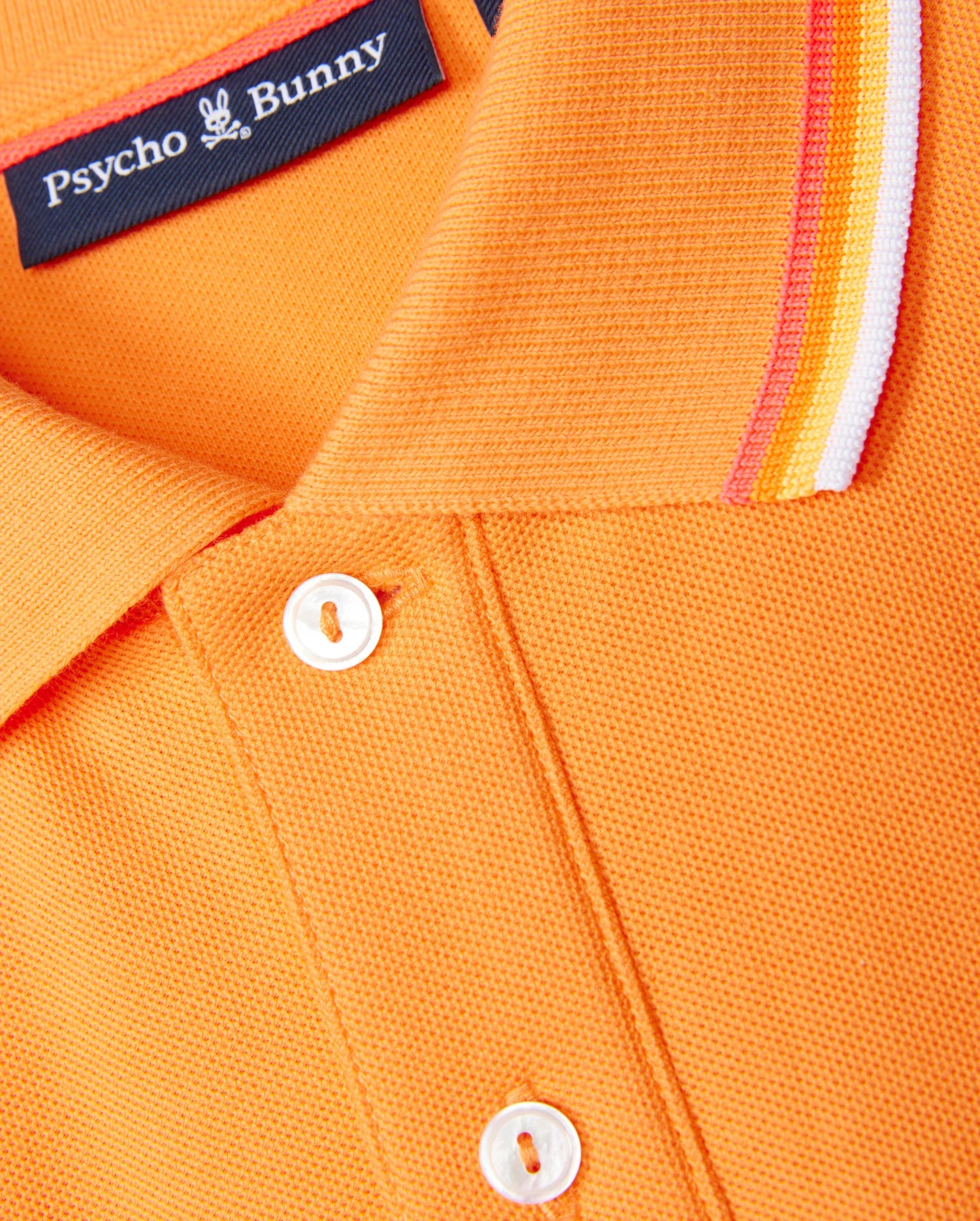 Close-up of a MENS GRANBURY PIQUE POLO - B6K400C200 in orange, showcasing a tipping collar with red, white, and yellow stripes. Crafted from diamond-knit Pima cotton, this shirt features white buttons and a navy tag bearing the "Psycho Bunny" brand name alongside a small bunny logo.