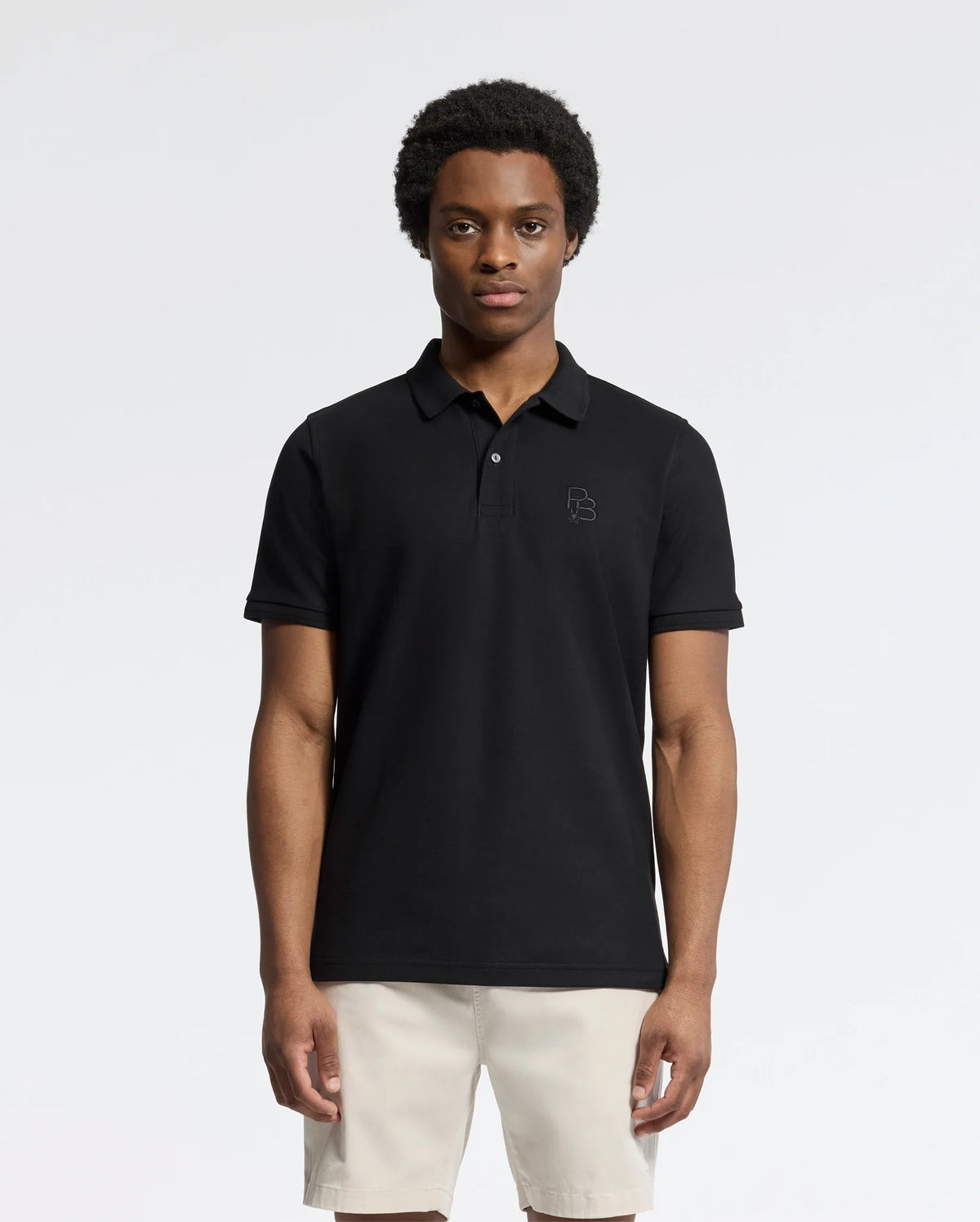 An individual in a Psycho Bunny MENS NICHOLSON RIB COLLAR PIQUE POLO (B6K401F200) in classic black and light-colored shorts stands against a white backdrop. This Pima cotton polo showcases an embroidered chest logo. The person has short curly hair and a neutral expression.